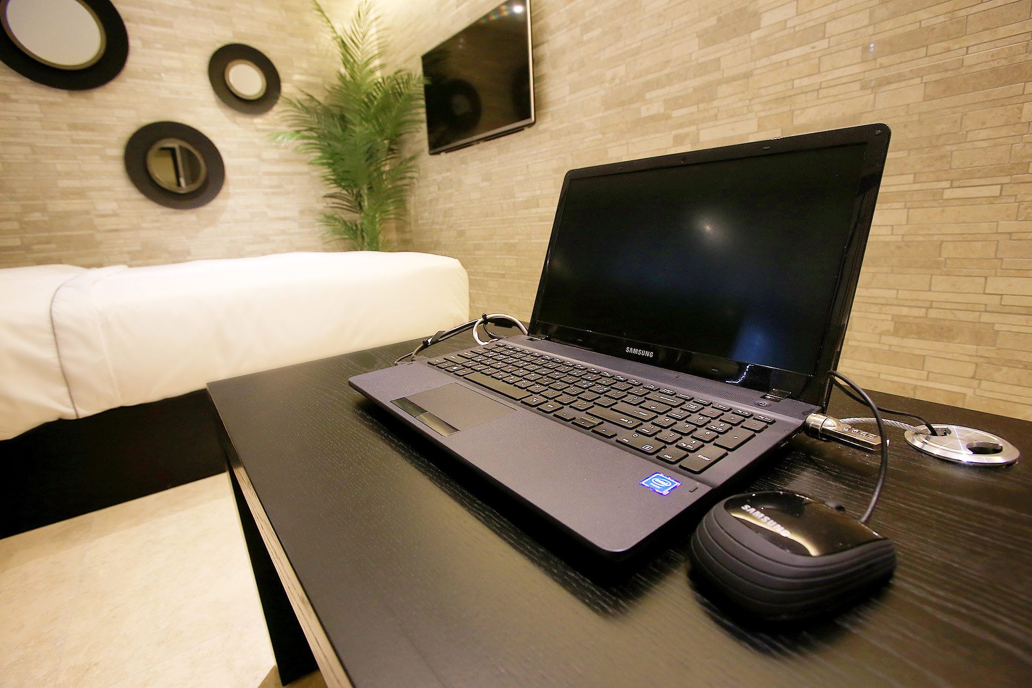 A laptop on a desk with a mouse, a plant, and a TV on the wall.