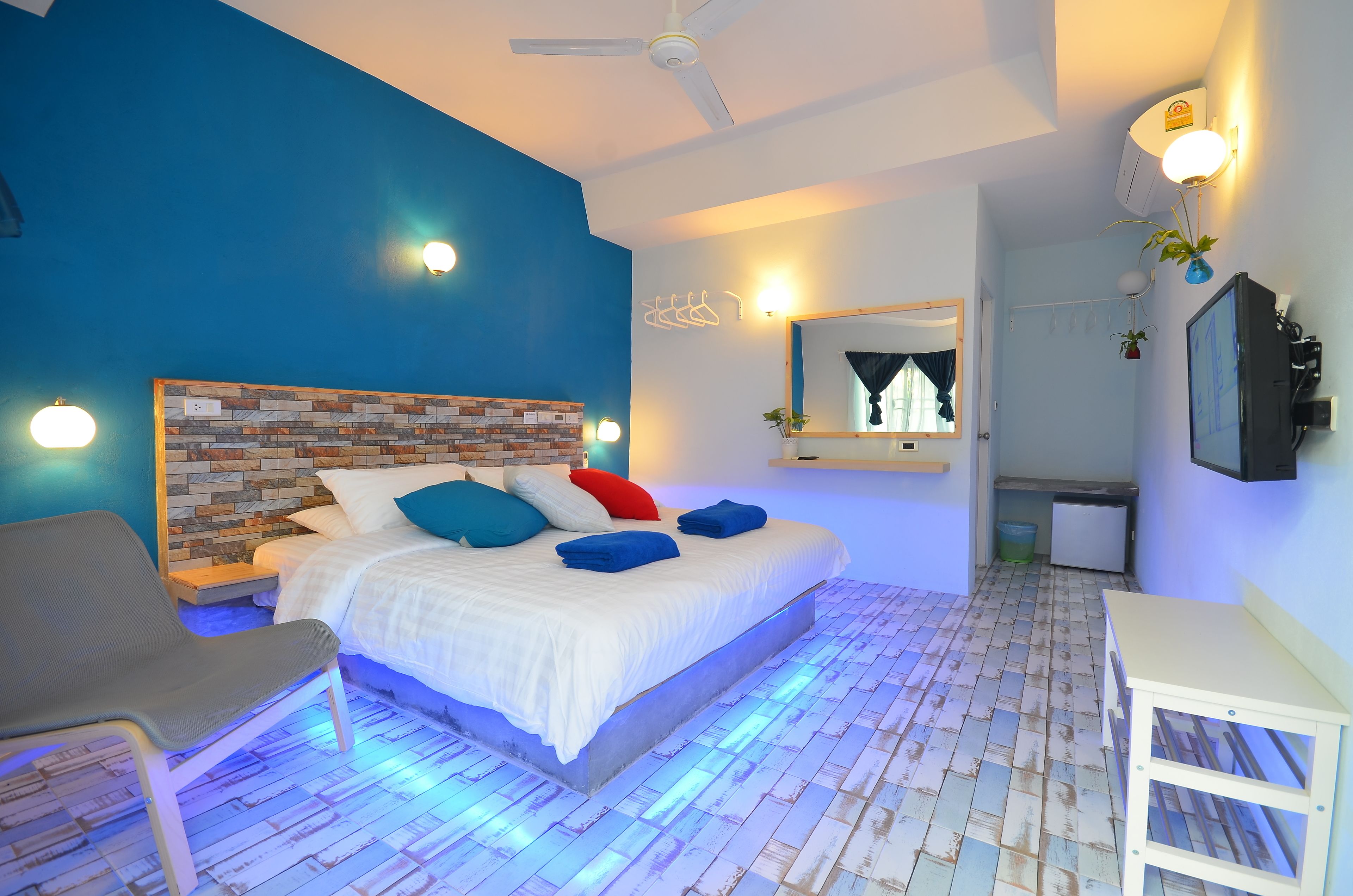Un dormitorio moderno con una pared azul decorativa, un cabecero con diseño de ladrillo, una cama con ropa de cama blanca y almohadas de colores, una silla gris y una mesita de noche pequeña.