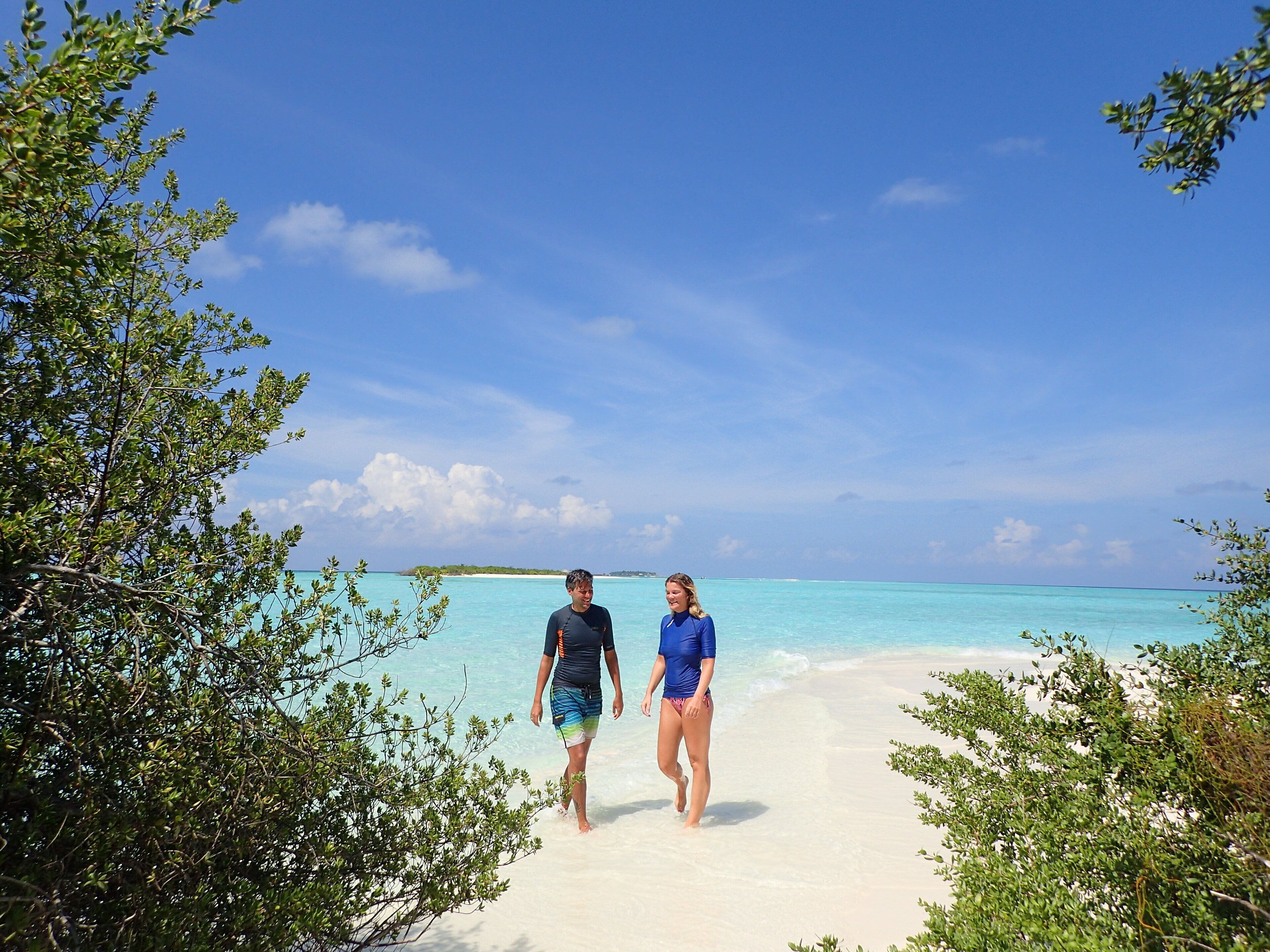 Two people walking on a sandy beach with clear turquoise water and a distant island.