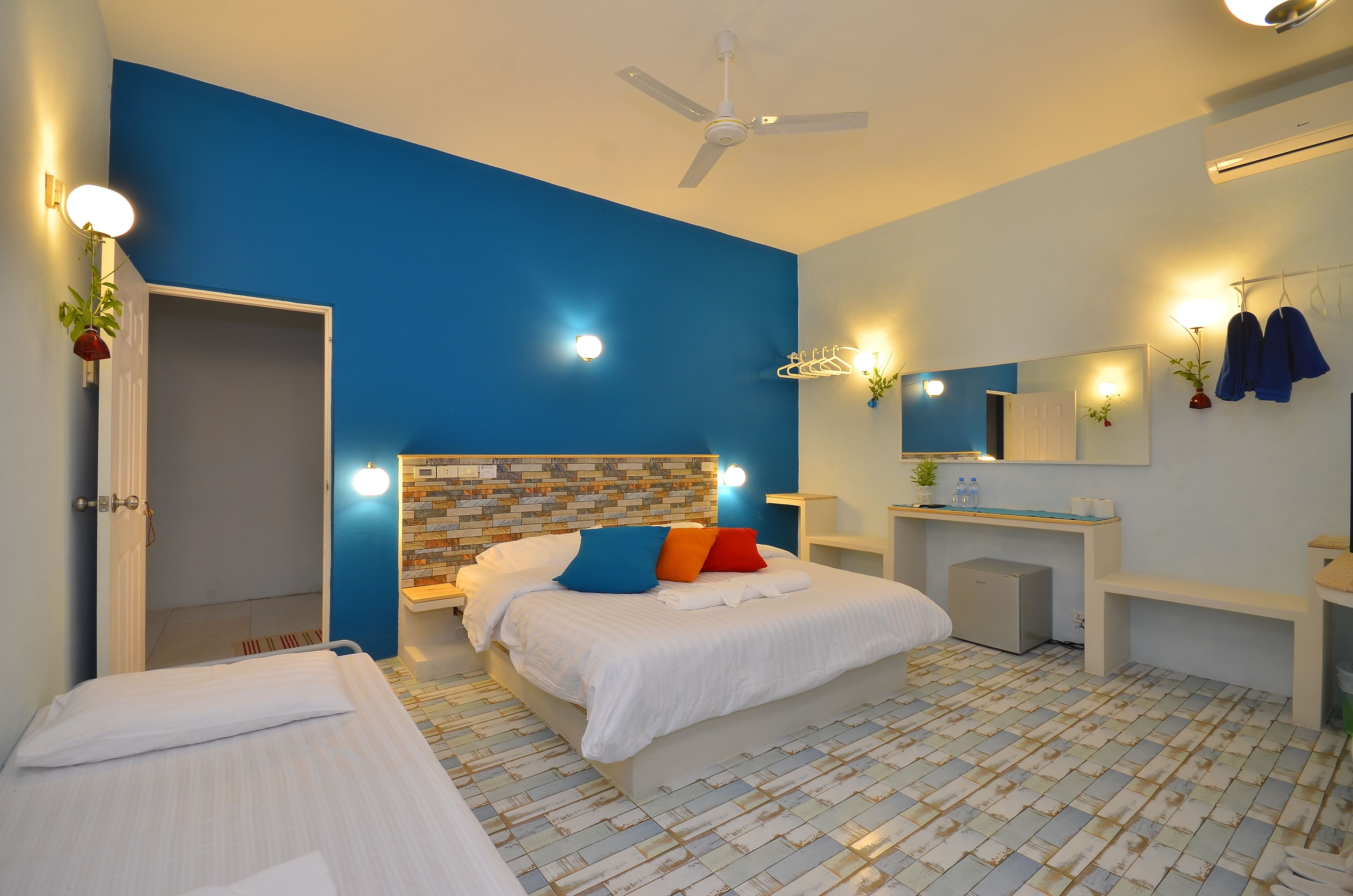 A modern bedroom with a blue accent wall, a white bed with colorful pillows, a ceiling fan, and a small bedside table with a plant.