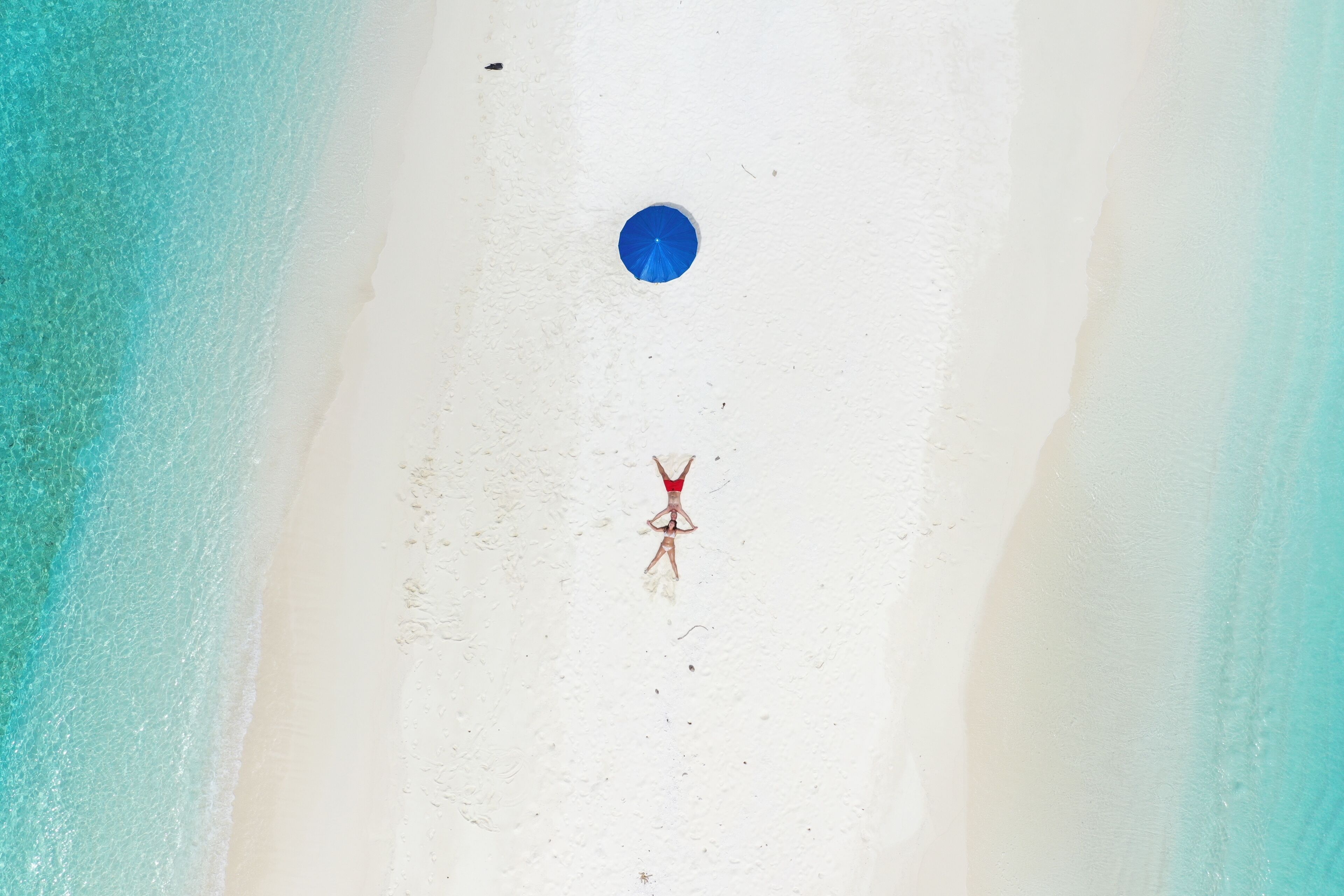 An aerial view of a beach with a person standing in the center, a blue object on the sand, and a clear demarcation between the sand and the water.