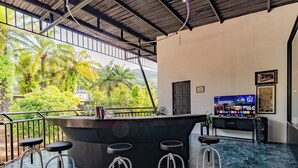 A bar area with stools, a TV displaying a show, and a view of greenery outside. - Villa Nap Dau 10 Bedrooms (Chalong)