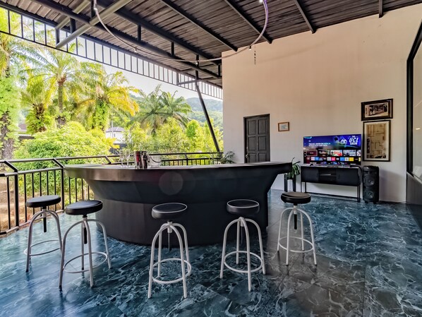 Zona de bar con taburetes, un televisor mostrando un programa y vistas al verdor exterior.
