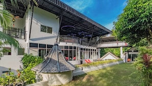 A modern two-story building with a flat roof, large windows, and a covered outdoor area with red chairs. - Villa Nap Dau 10 Bedrooms (Chalong)