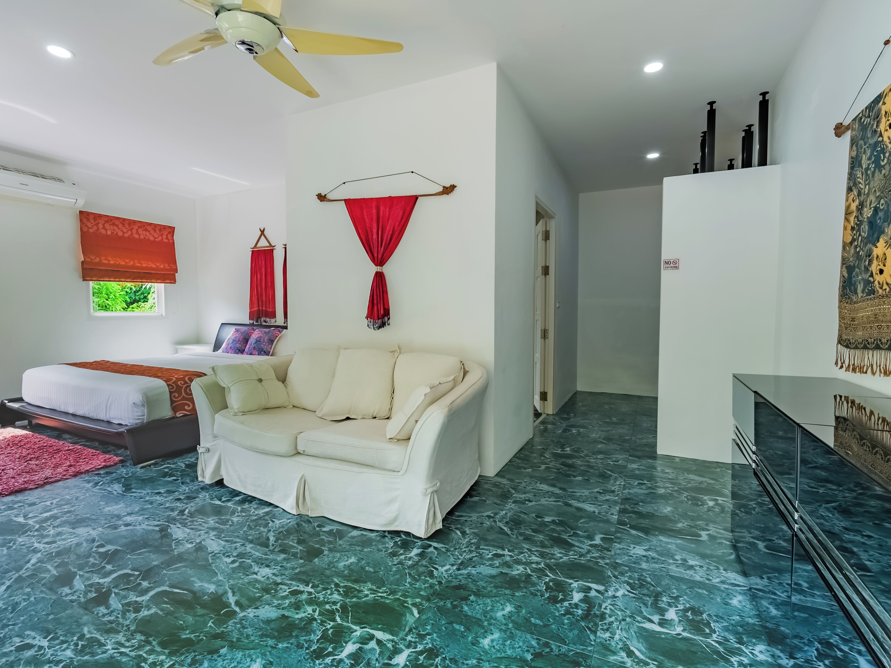 A hotel room with a bed, a white sofa, a red curtain, a ceiling fan, and a marble floor.