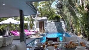 En matplats vid poolkanten med ett glasbord, vita och lila loungestolar och utsikt över en swimmingpool.