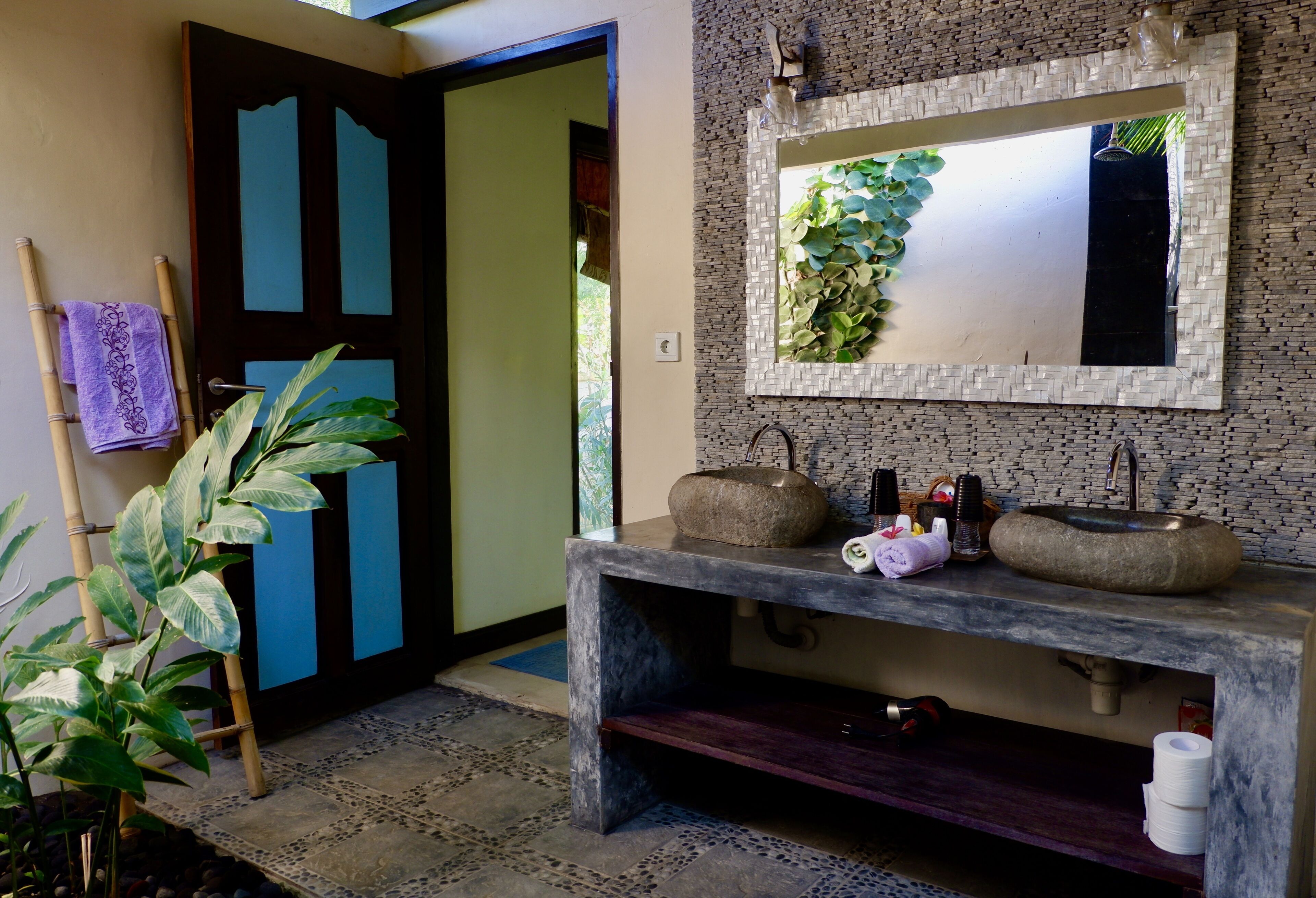 A bathroom with a stone sink, a mirror with a plant, and a bamboo towel rack.