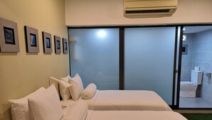 A hotel room with two beds, a large window, and a bathroom visible through the glass door.