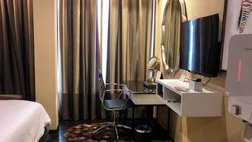 Habitación de hotel con cama, escritorio, silla, televisor y cortinas.