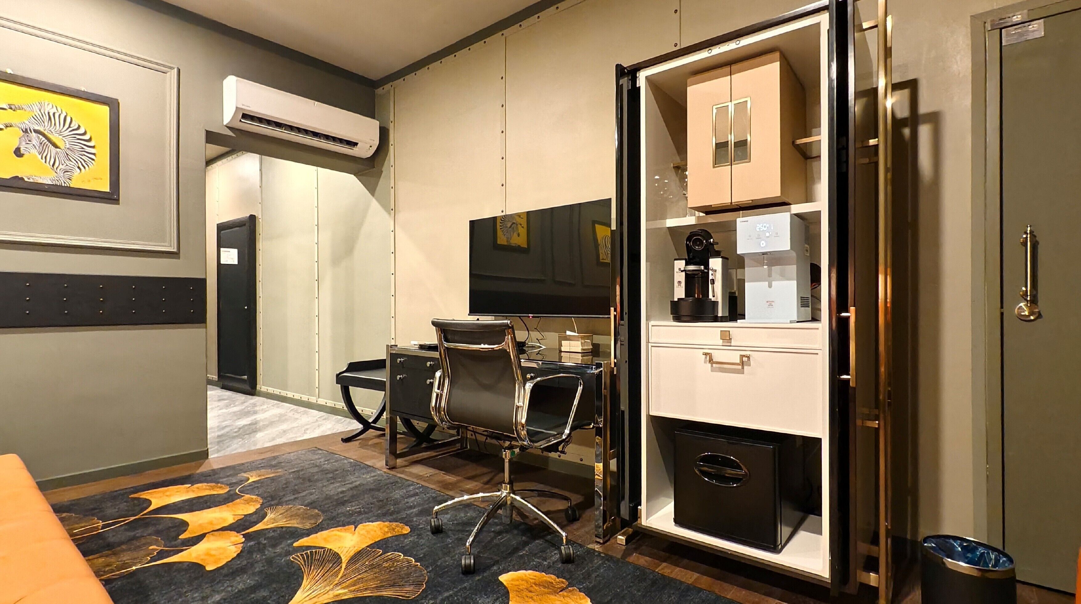 A hotel room with a desk, chairs, a coffee machine, a TV, and a minibar.