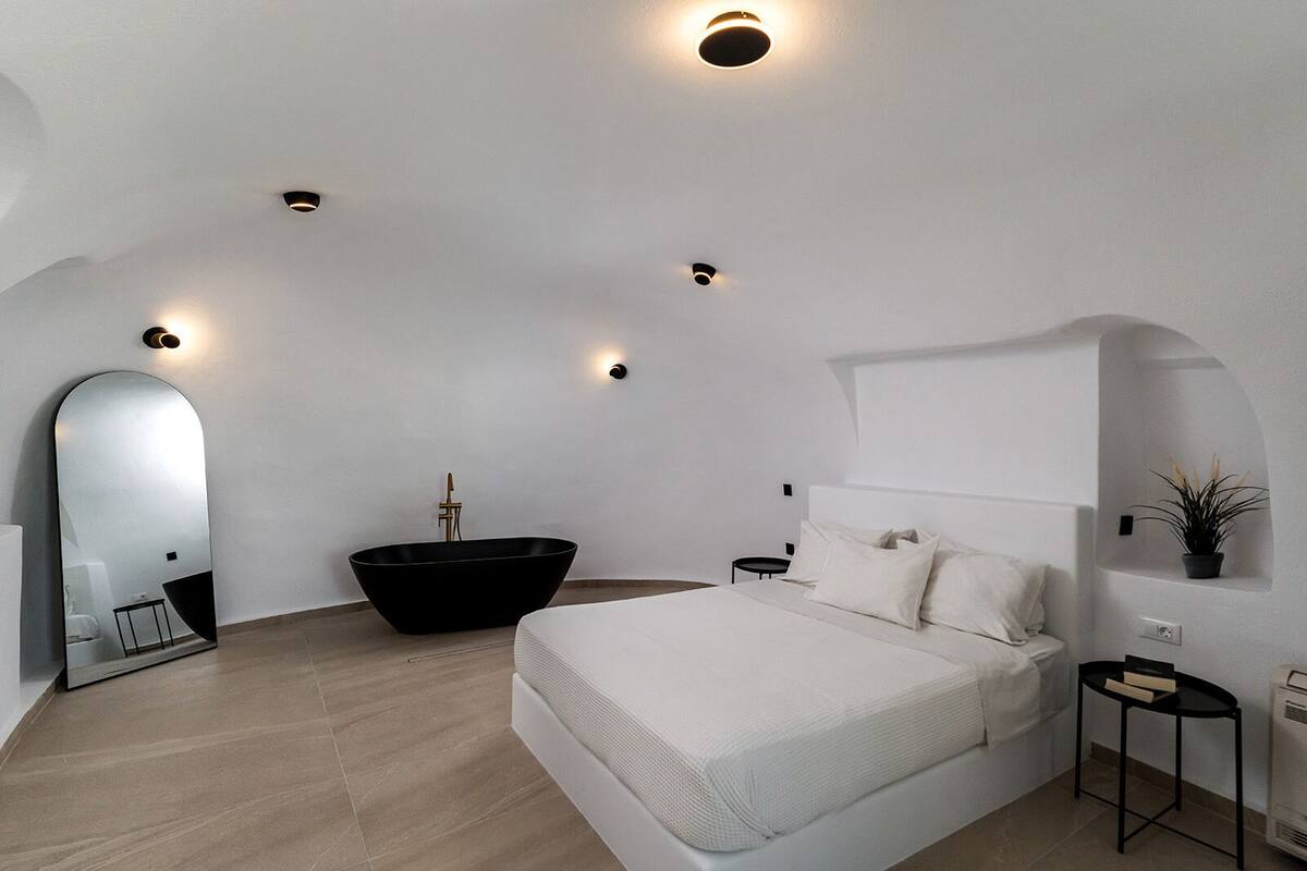 A modern bedroom with a large bed, a black freestanding bathtub, a round mirror, and recessed ceiling lights.