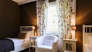 Une chambre avec deux lits, un fauteuil blanc, une table de chevet avec une lampe et une fenêtre ornée de rideaux à motifs de feuilles.