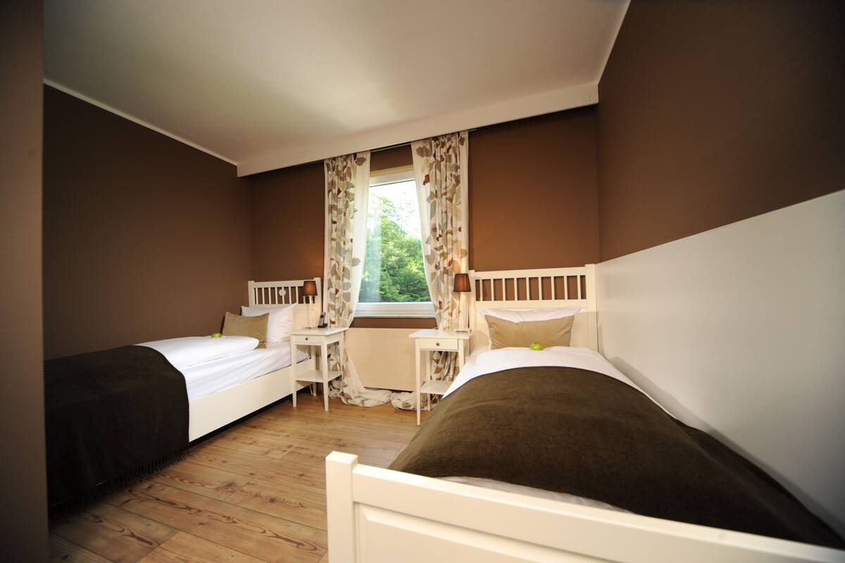 A bedroom with two single beds, a small table, a chair, and a window with curtains.