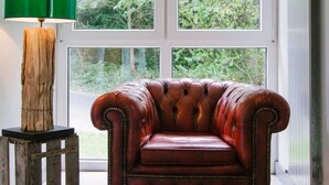 Une pièce comprenant un fauteuil en cuir, une table d’appoint en bois, un lampadaire vert et une grande fenêtre donnant sur un paysage verdoyant.