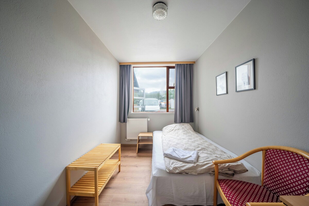 Una habitación pequeña y bien iluminada con una cama individual, un banco de madera, un radiador y una ventana con cortinas.