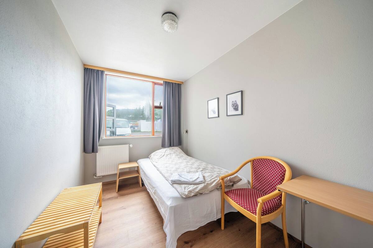 A small, well-lit room with a bed, a desk, a chair, and a window with curtains.