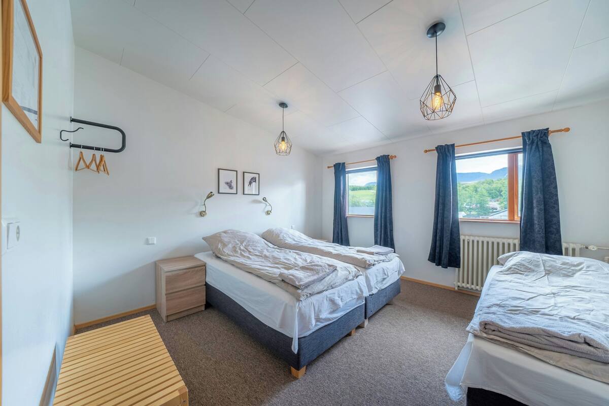 A bedroom with two beds, a wooden bench, a radiator, and a window with curtains.