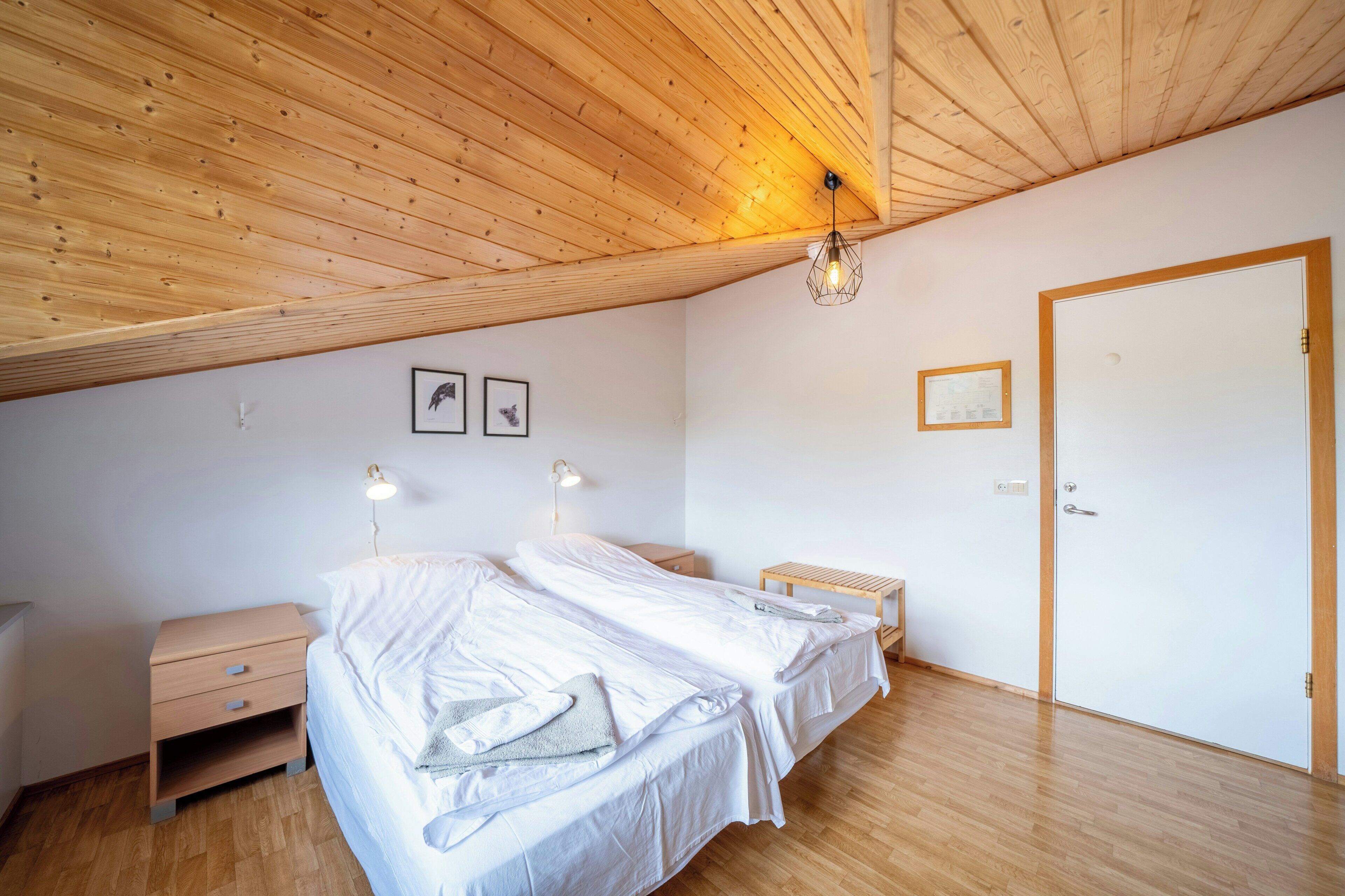 A bedroom with two beds, wooden flooring, a wooden ceiling, a door, and wall-mounted lights.
