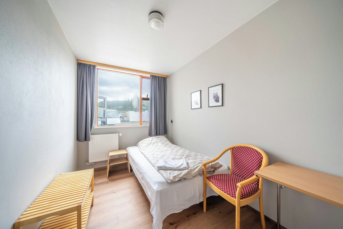 A small, well-lit room with a bed, a desk, a chair, and a window with curtains.
