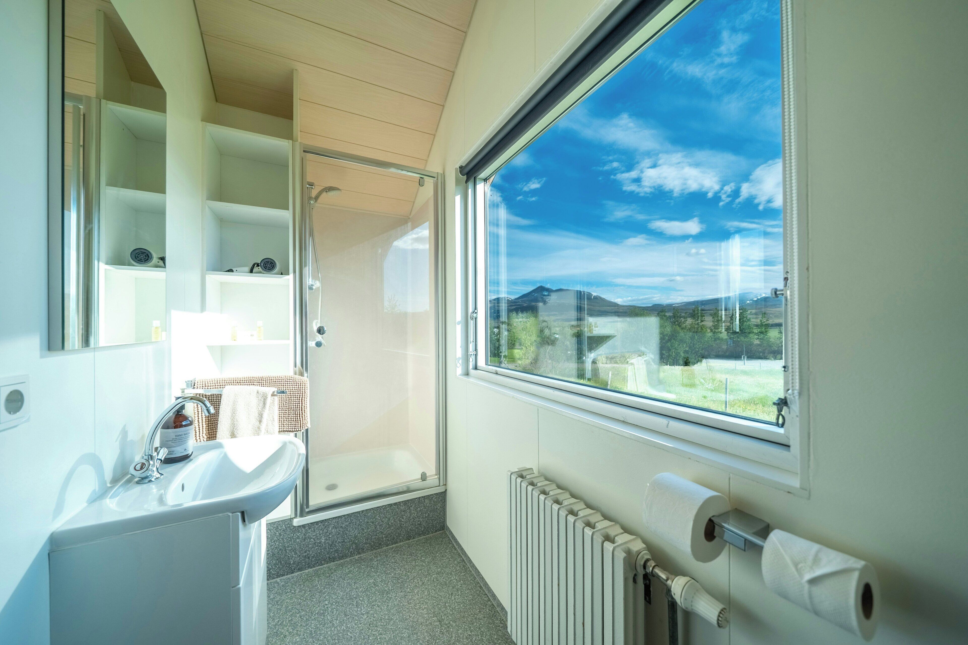 Een badkamer met een wastafel, spiegel, douche en toilet. Er is een groot raam met uitzicht op de omgeving.