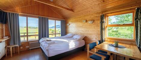 A wooden cabin room with a bed, a table with chairs, a desk, and large windows offering a view of the outdoors.