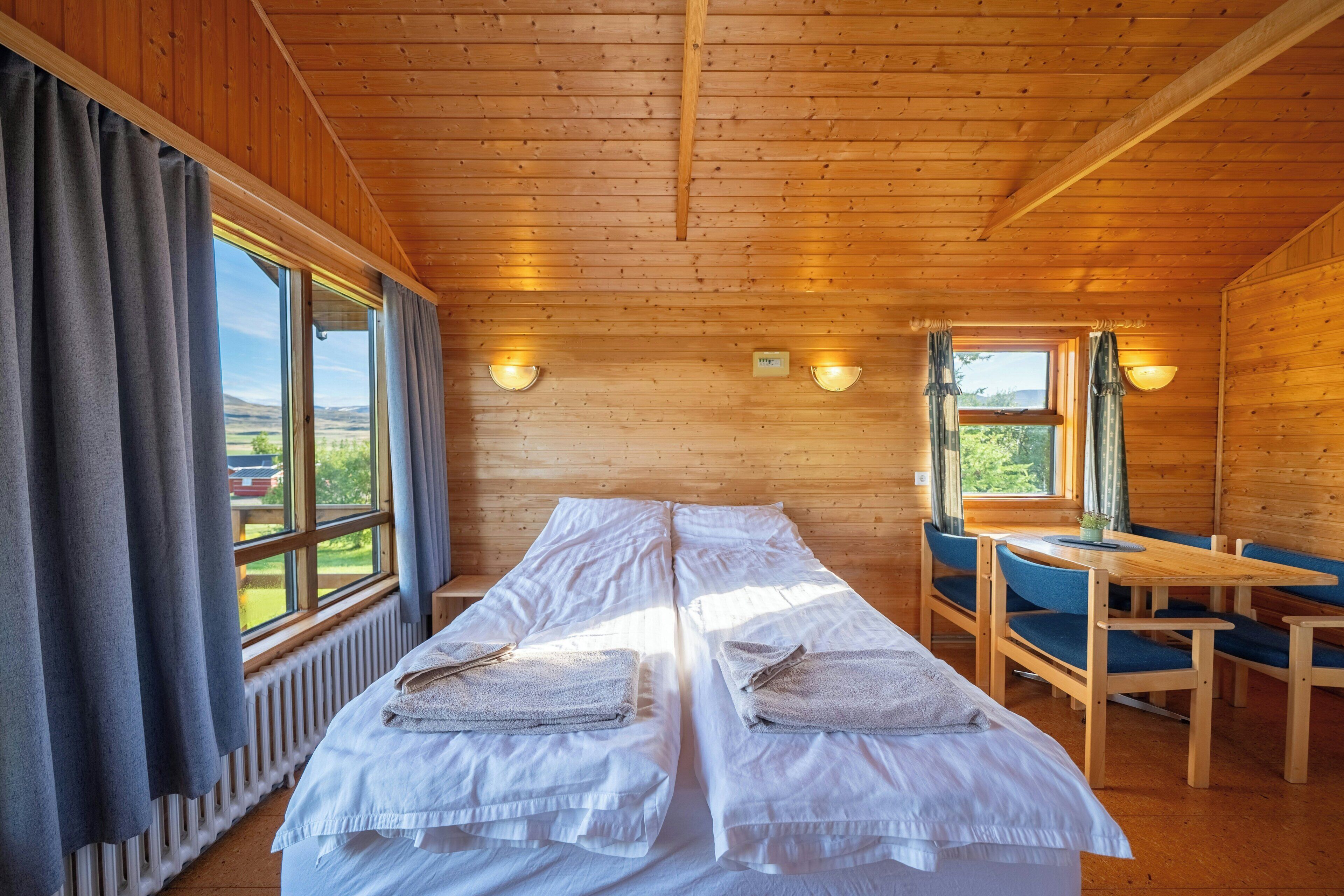 Quarto de cabana de madeira com duas camas, uma mesa, cadeiras e janelas amplas com cortinas.