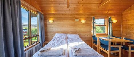 A wooden cabin room with two beds, a table, chairs, and large windows with curtains.