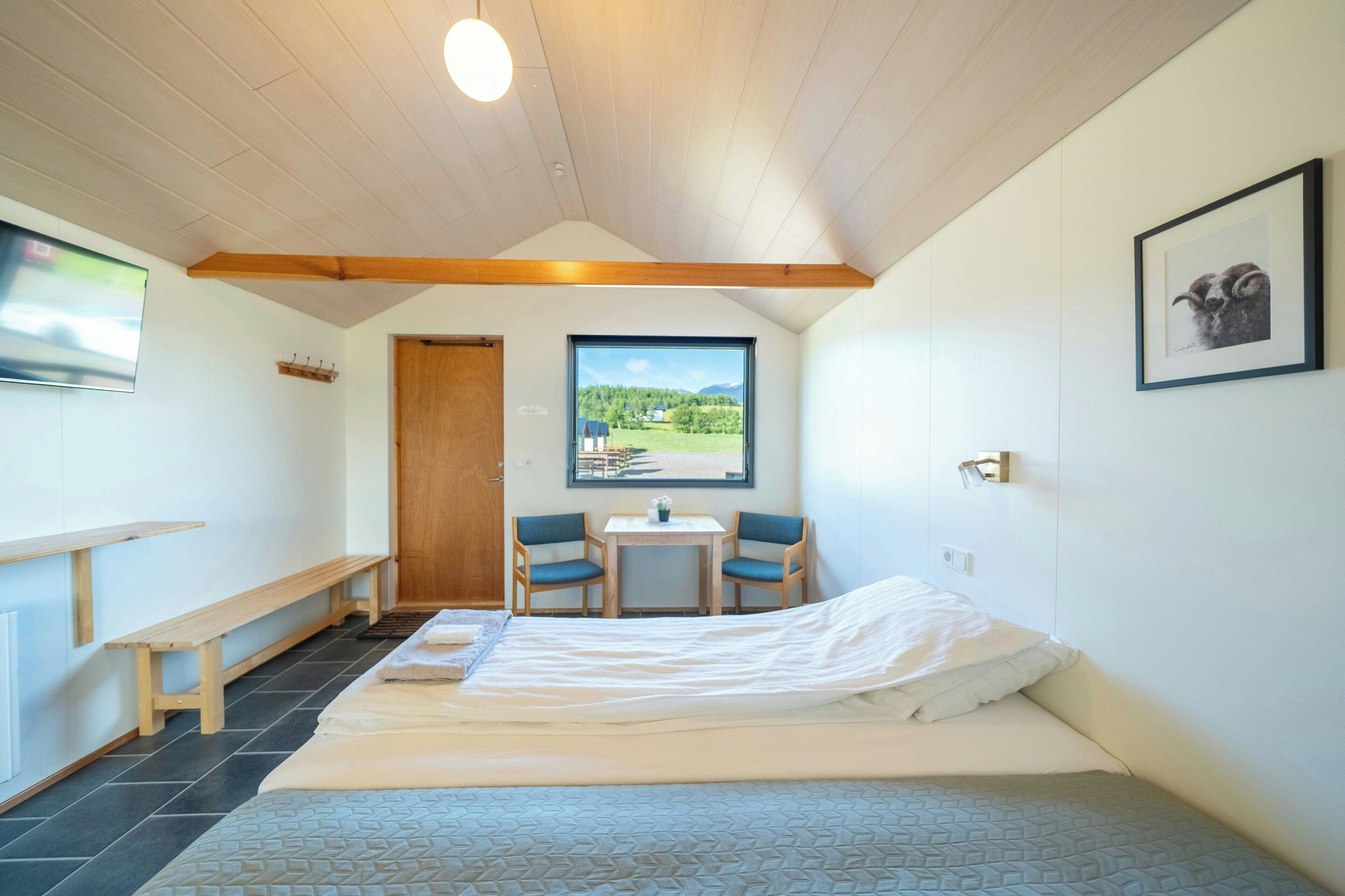 Un dormitorio con cama, una mesita, dos sillas, un banco, ventana con vistas y un cuadro en la pared.