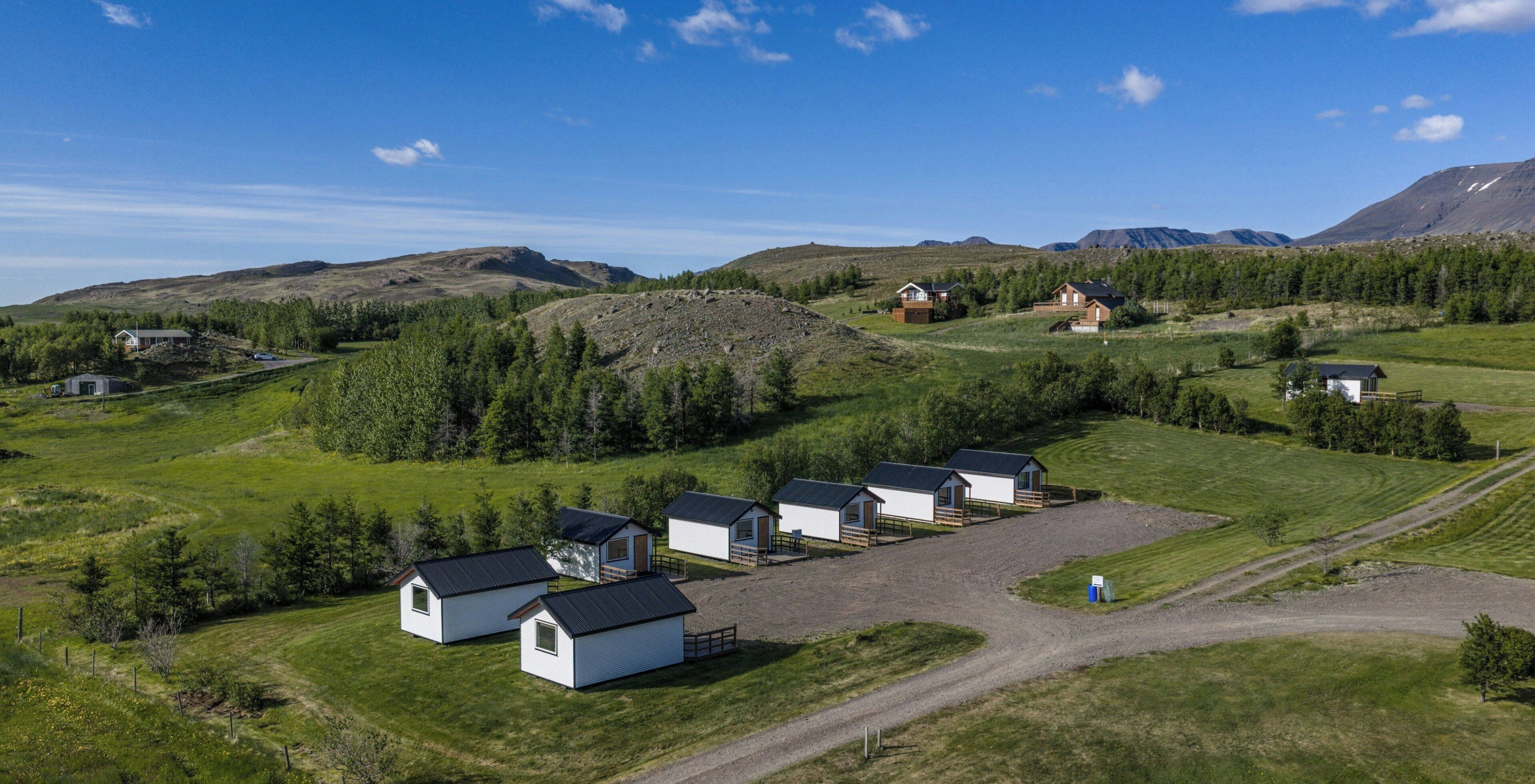 A rural landscape with several small cabins and a larger house, surrounded by greenery and mountains.