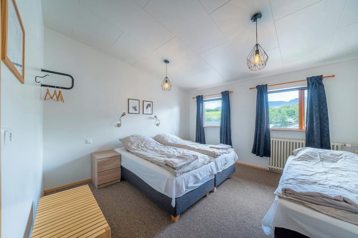 A bedroom with two beds, a wooden bench, a radiator, and a window with curtains.