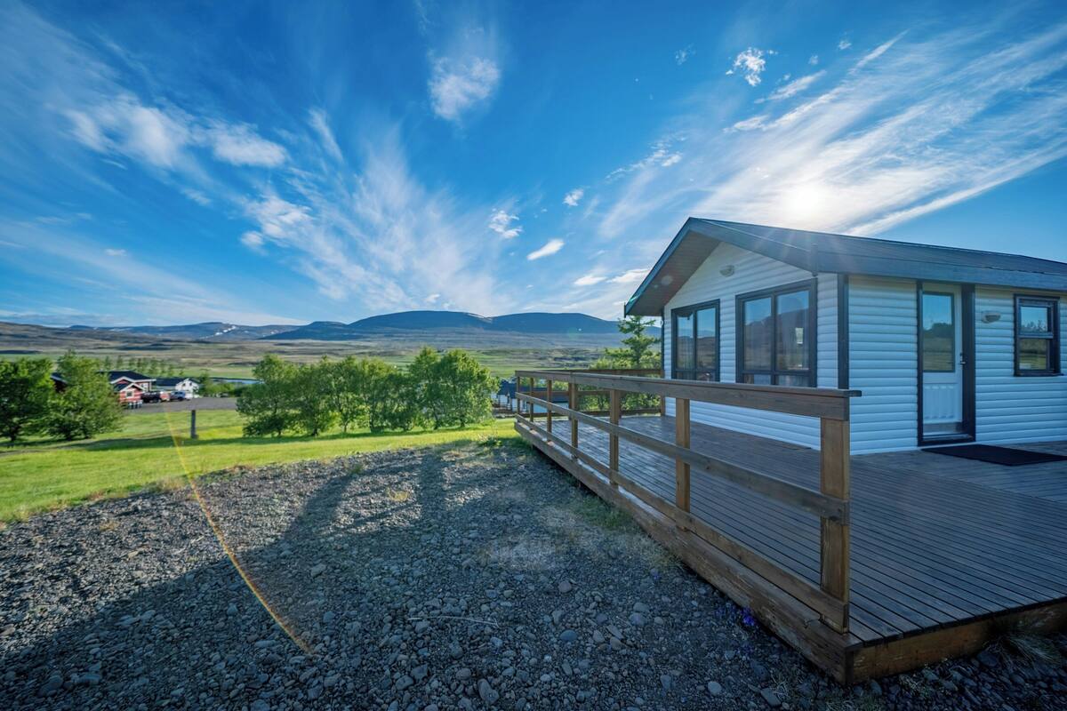 A modern cabin with a wooden deck, large windows, and a view of the surrounding landscape.