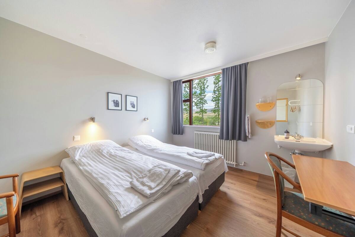 A bedroom with two beds, a window with curtains, a radiator, a sink, and a wooden table.