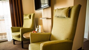 Une chambre d’hôtel avec deux fauteuils verts, une petite table sur laquelle se trouvent une bouteille et des fruits, et un téléviseur fixé au mur.