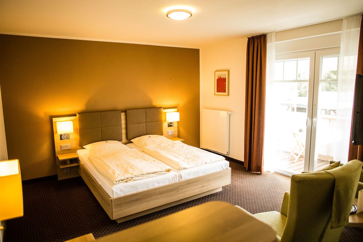 A hotel room with a double bed, two bedside tables with lamps, a large window with curtains, and a green armchair.