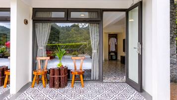 A hotel room with a sliding glass door leading to a balcony with a view of the mountains. There are two wooden chairs and a small table with a plant on the balcony.