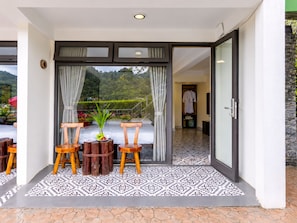 Habitación de hotel con puerta corrediza de vidrio que da a un balcón con vistas a las montañas. Hay dos sillas de madera y una mesita con una planta en el balcón.
