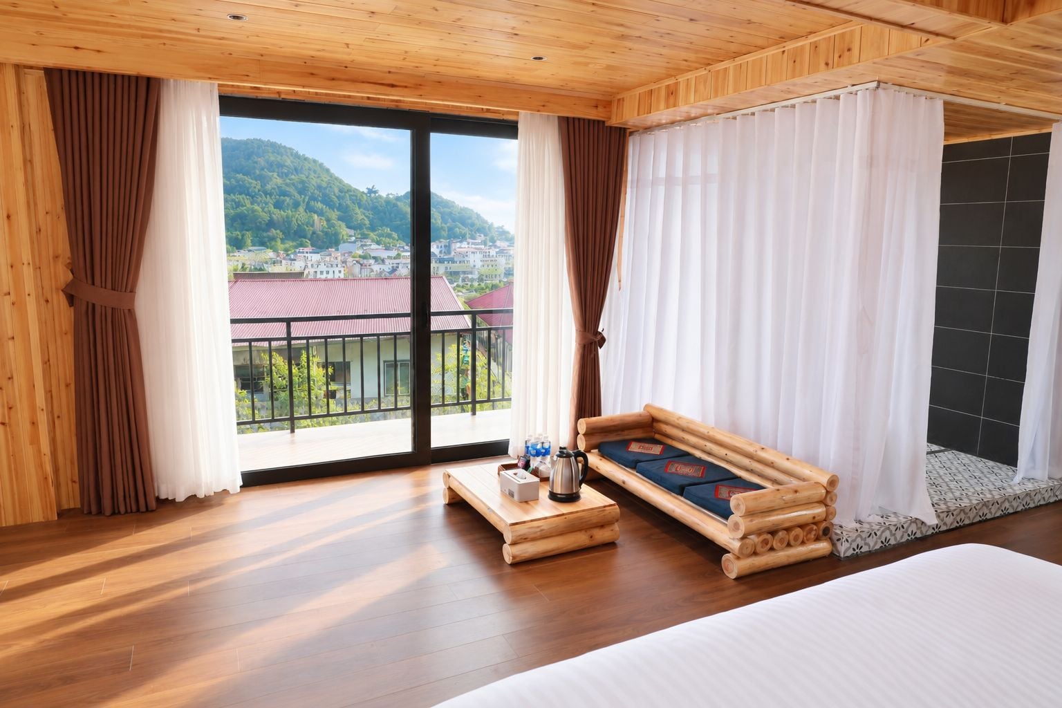 Habitación de hotel con un ventanal grande, suelo de madera, una cama, una mesita con una tetera y botellas, y un banco de madera con cojines.