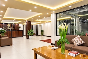 Zona de recepción de un hotel con mostrador, recepcionista y área de descanso con sofá y mesa de centro.