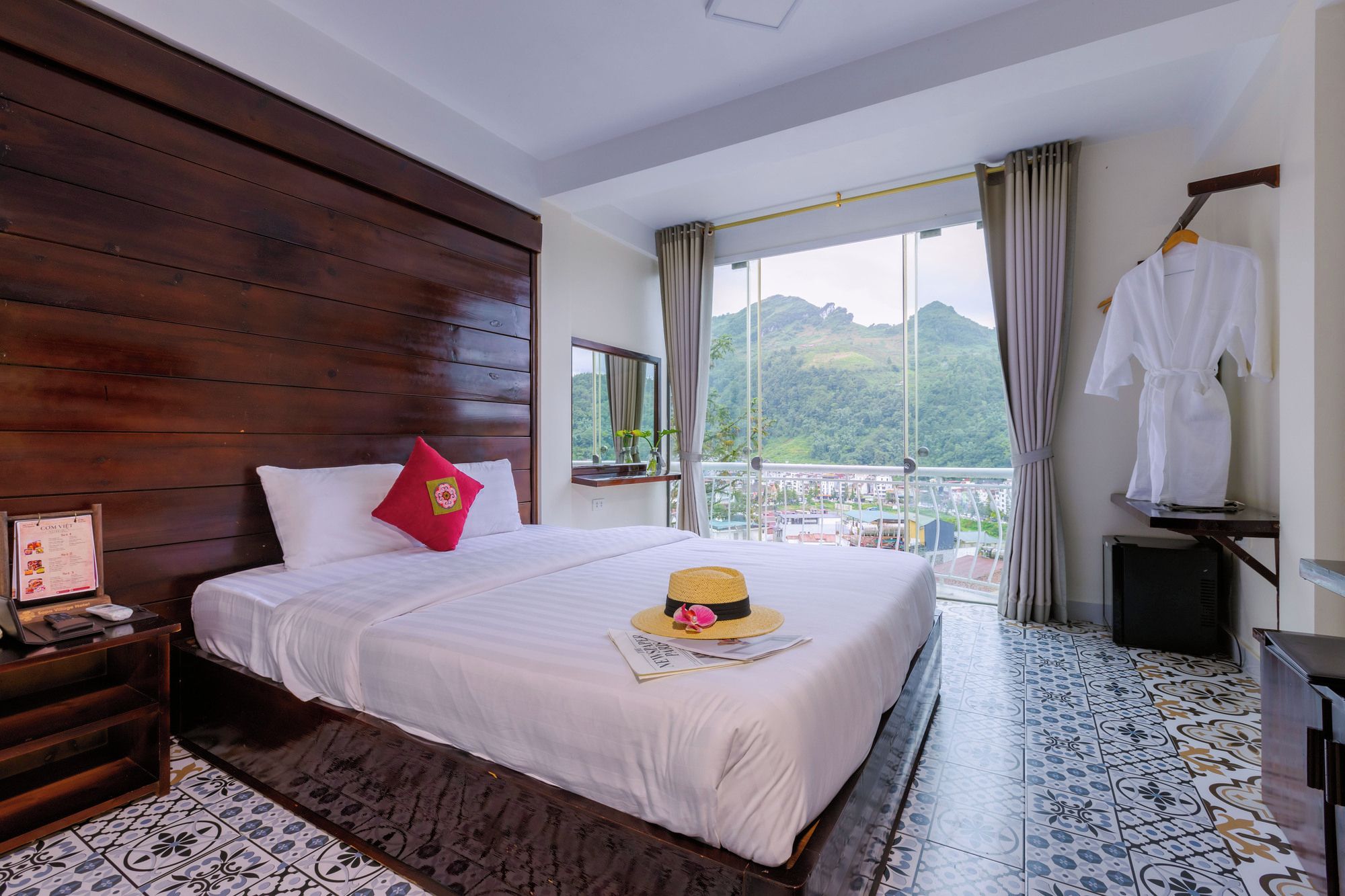 Una habitación de hotel con una cama grande, cabecero de madera, vistas a las montañas y un balcón con una bata blanca colgada.