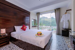 A hotel room with a large bed, a wooden headboard, a view of the mountains, and a balcony with a white robe hanging. - Sapa Village Hotel Sky Lounge & Spa (Sa Pa)