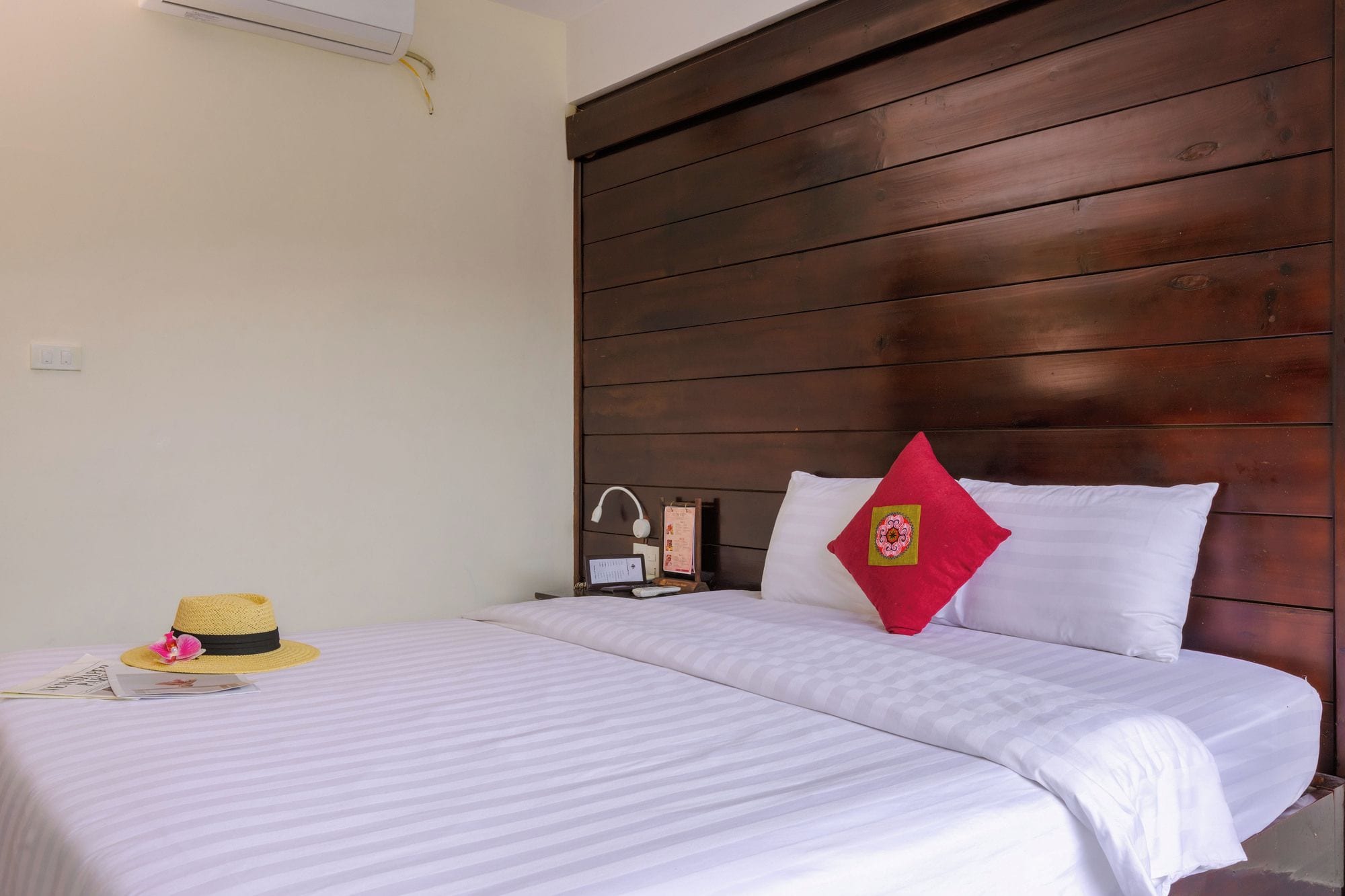 Habitación de hotel con cabecera de madera, cama con ropa blanca, almohada roja, sombrero de paja y escritorio con lámpara y computadora.
