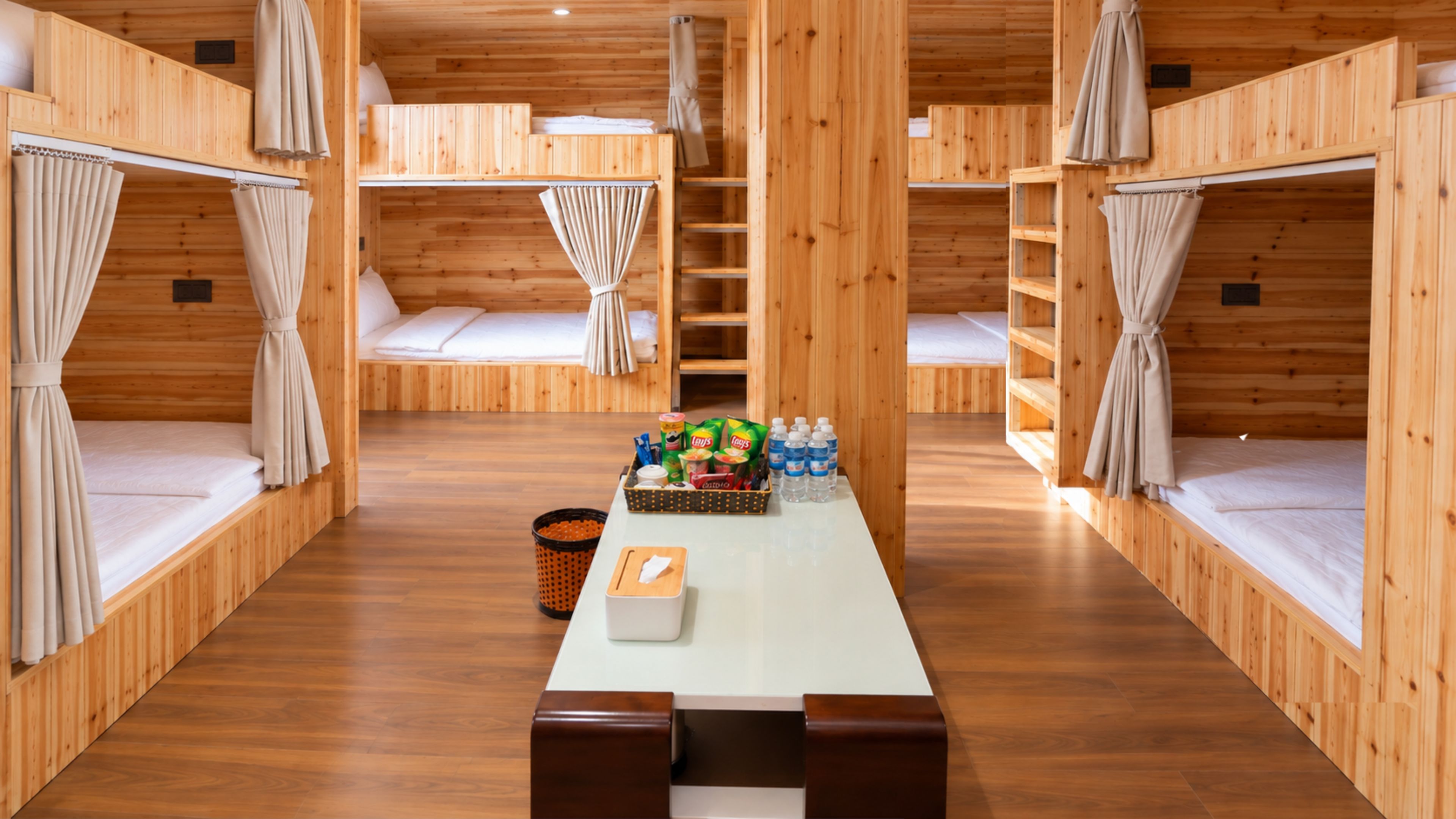 Una cabaña de madera con literas, una mesa central con aperitivos y un pequeño cubo de basura.