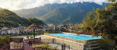 Una piscina con borde de piedra, rodeada por una escalera de piedra y una barandilla de metal, con vistas a un pueblo y montañas.