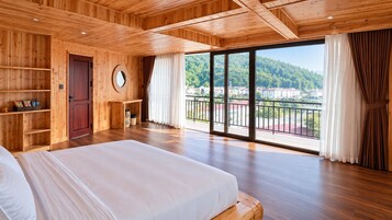 Un dormitorio amplio con una cama grande, paredes de madera, techo de madera, un balcón con vista a un pueblo y colinas, y un escritorio de madera con una silla.