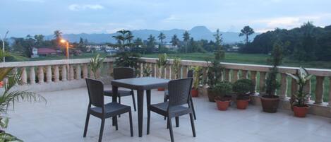Una terraza con mesa y sillas, rodeada de plantas en macetas, con vistas a un paisaje verde y montañas al horizonte.
