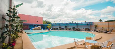 Una piscina con sillones de descanso y mesas, rodeada de plantas en macetas y un edificio al fondo.