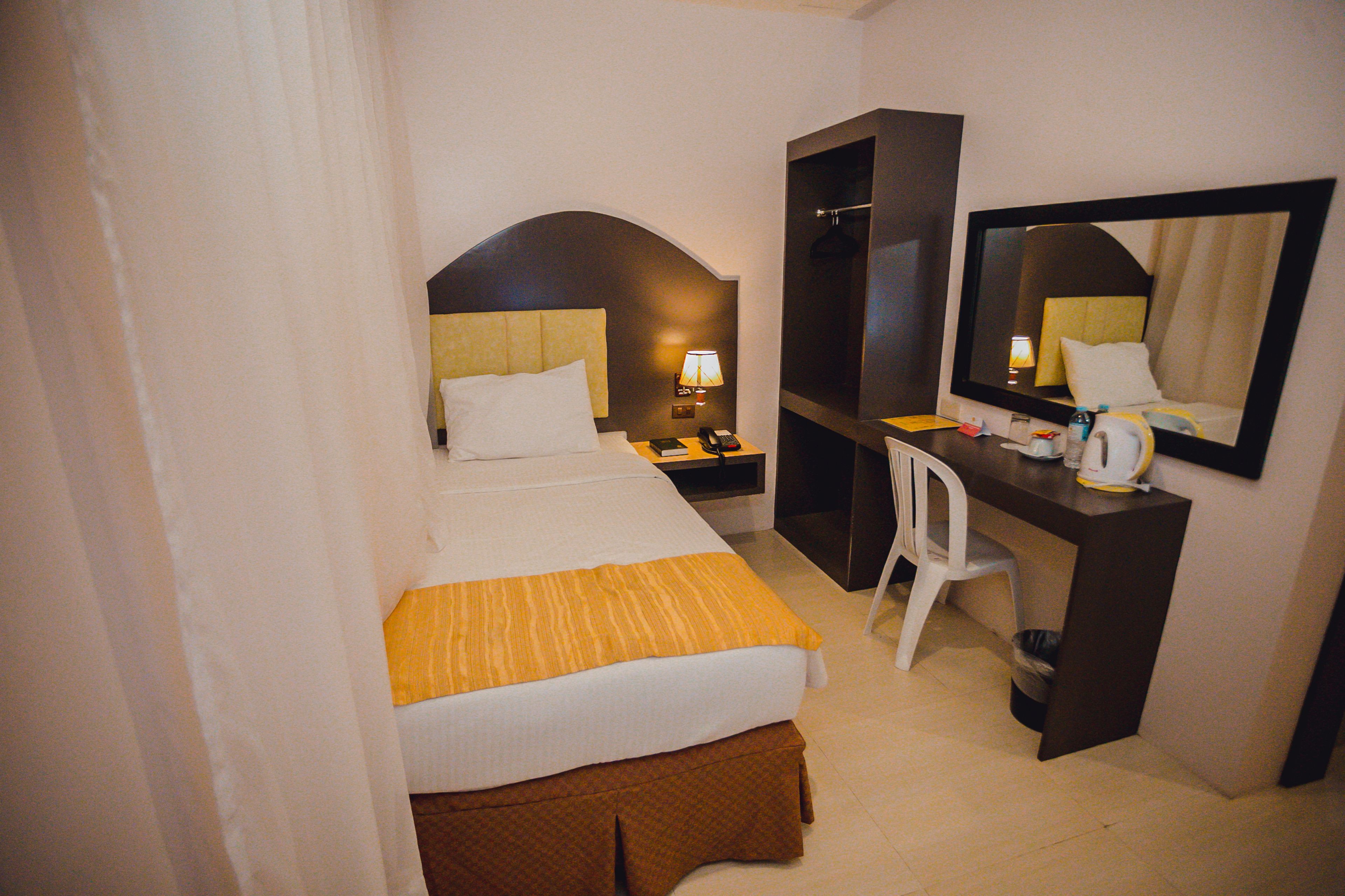 A hotel room with a bed, a desk, a chair, a mirror, and a nightstand with a lamp.