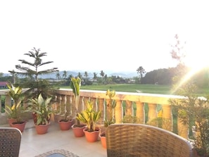 Uma varanda com vasos de plantas, cadeiras de vime e vista para uma paisagem verde com árvores e montanhas ao longe.
