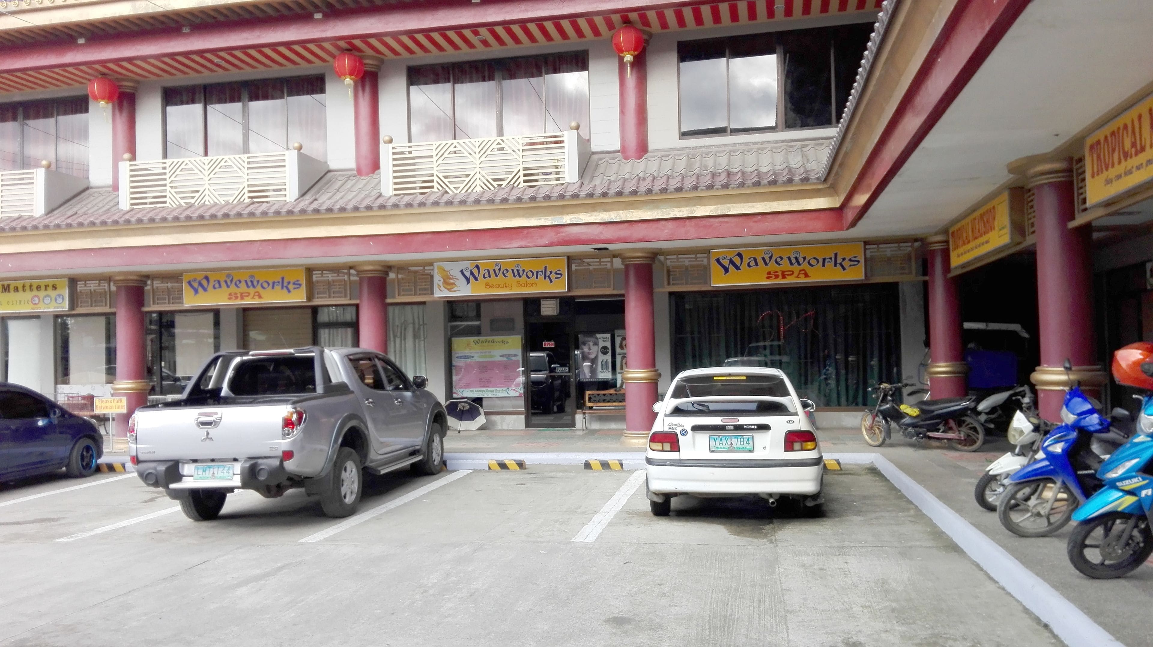 A building with multiple businesses including a beauty salon and a spa. There are several parked vehicles and motorcycles in front.