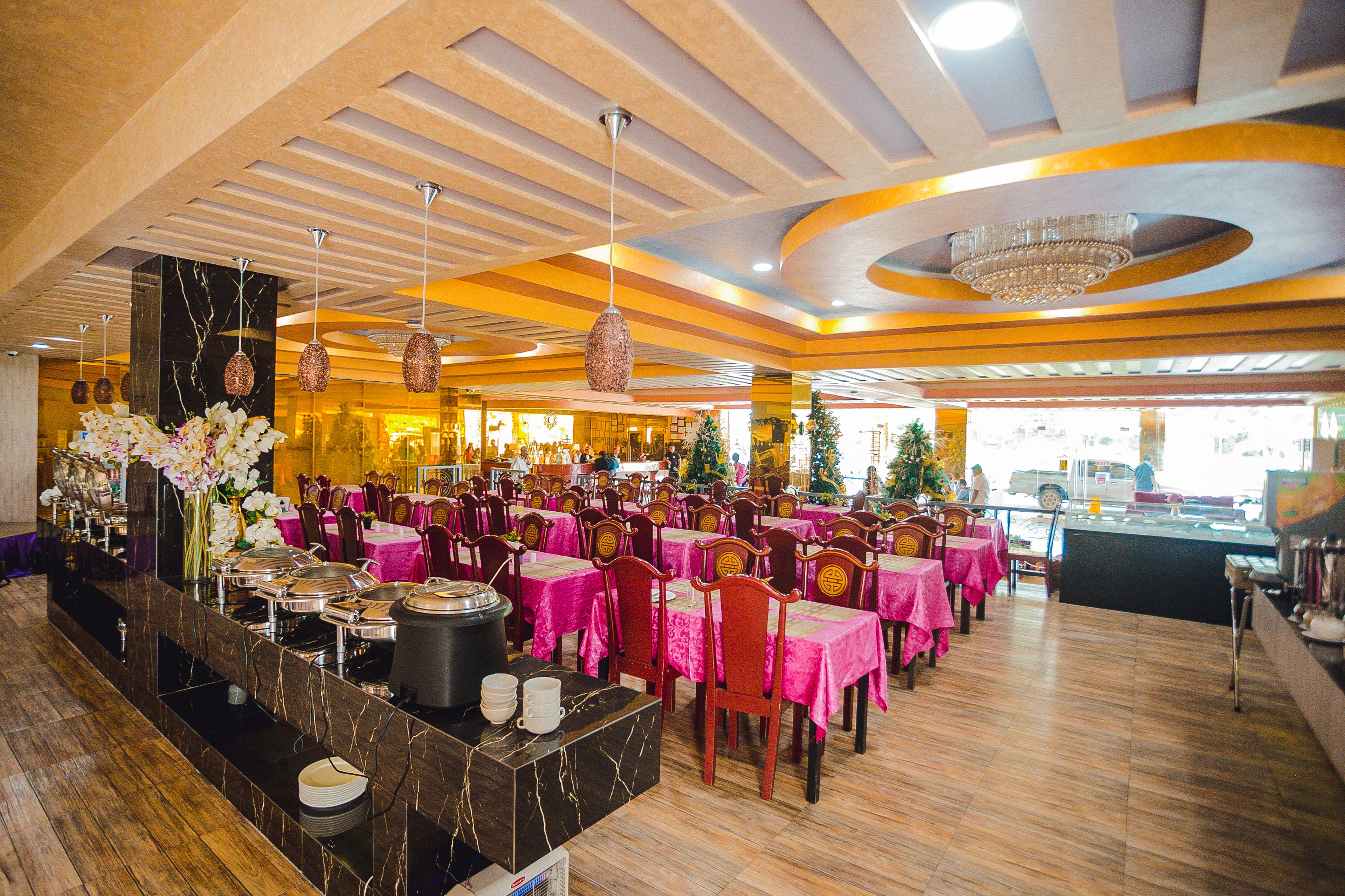 A restaurant with multiple tables covered in pink tablecloths, a buffet area with various dishes, and a chandelier on the ceiling.